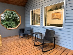 A covered porch with two wicker chairs and a small table, wooden flooring, and a view through a window.