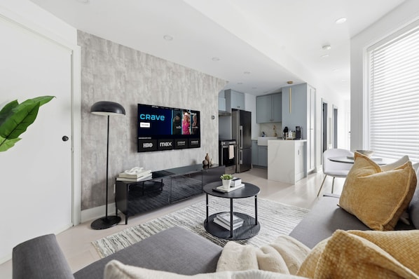 A modern living room with a flat-screen TV, a sofa, a coffee table, and a kitchen in the background. - Stylish 2BR Retreat | Private Patio & Free Park (Toronto)