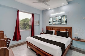 Una habitación de hotel con cama, mesita de noche, teléfono, ventana con cortinas, equipo de aire acondicionado y obra de arte colgada en la pared.