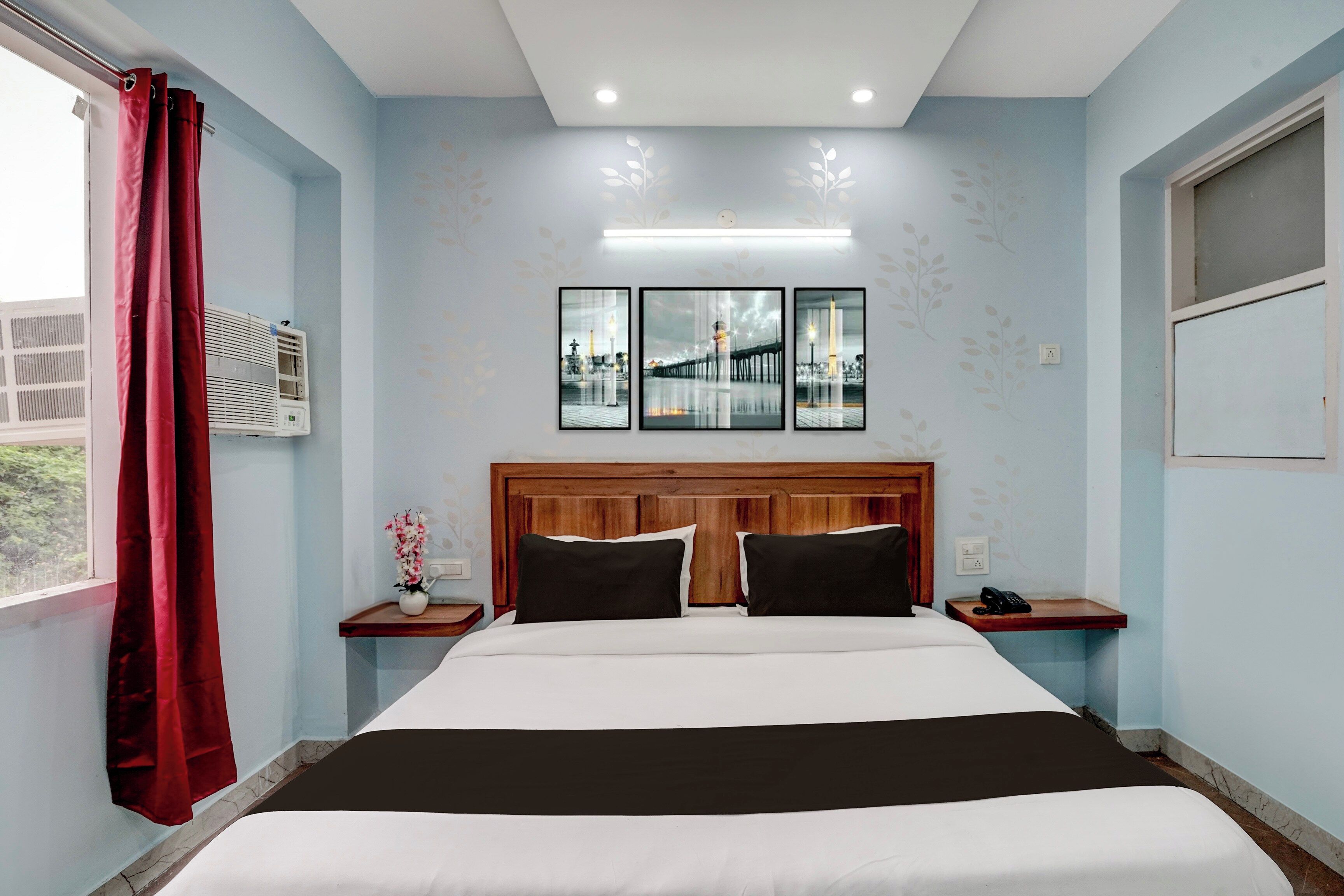 Habitación de hotel con cama, mesita de noche, aire acondicionado de pared, ventana con cortinas rojas y pared decorada con tres cuadros.