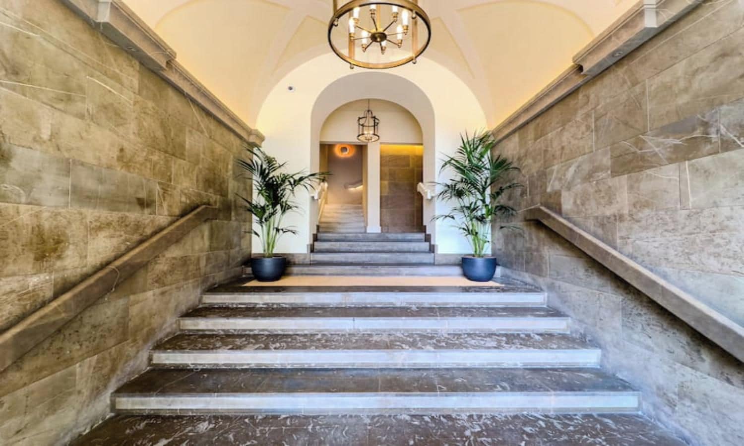 A grand staircase with marble steps, flanked by stone walls, leading to a room with a chandelier.