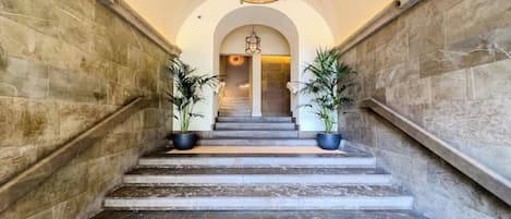 A grand staircase with marble steps, flanked by stone walls, leading to a room with a chandelier.