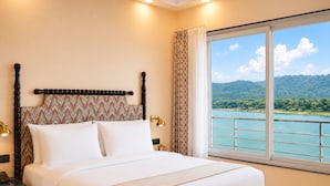 Ein Hotelzimmer mit Bett, Nachttisch, Lampe und Fenster mit Blick auf einen See und Berge.