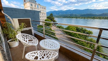 A balcony with two white chairs and a potted plant, overlooking a river and mountains.