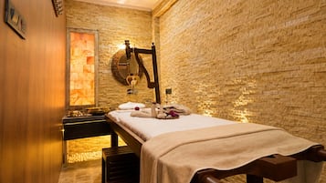 Un cuarto de spa con una camilla de masaje, un banco de madera, una pared de piedra y un espejo.