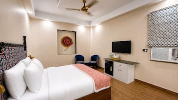 Quarto de hotel com cama, escrivaninha, cadeira, ventoinha no teto, televisão e aparelho de ar condicionado.
