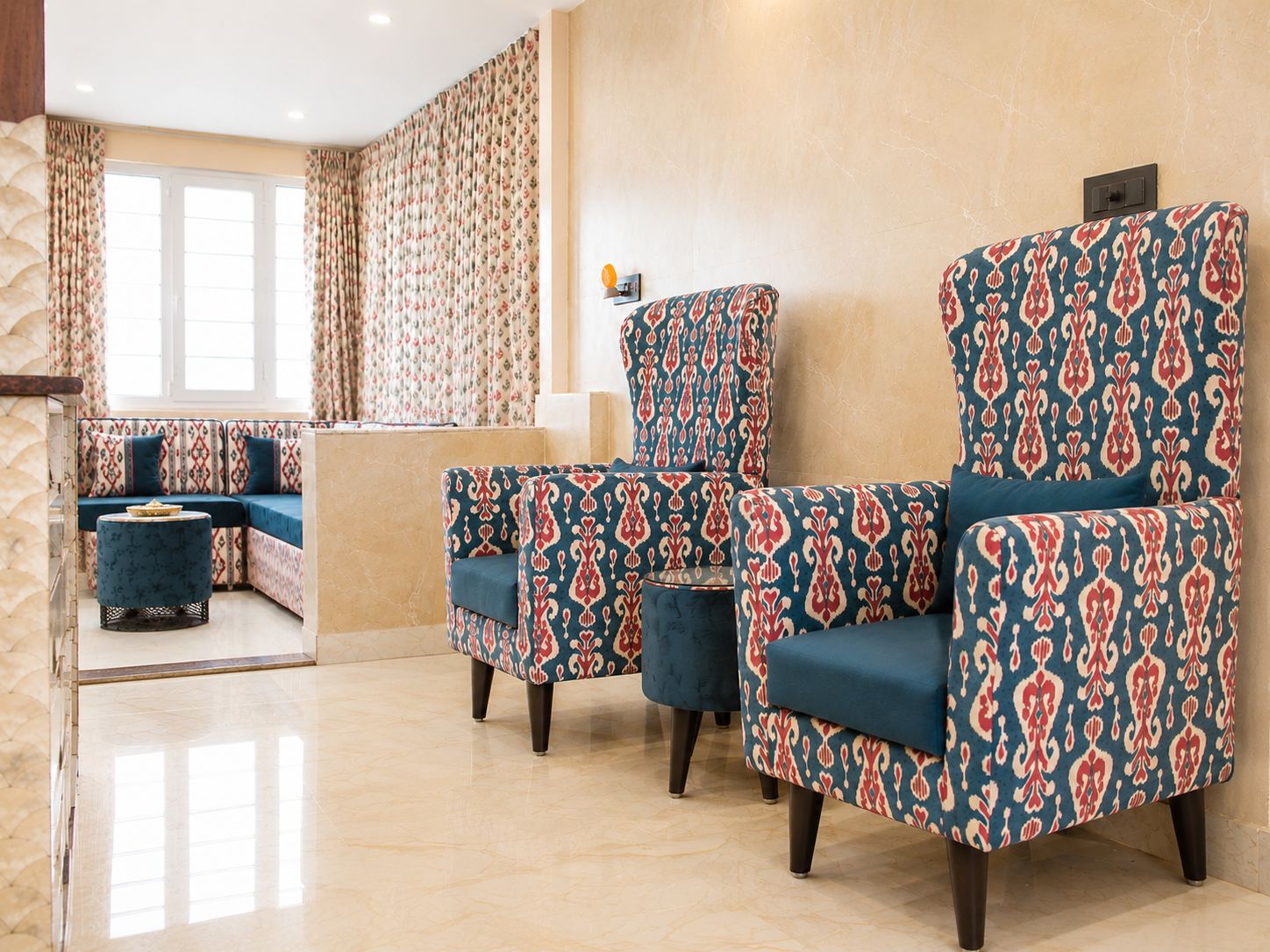 A hotel room with a seating area featuring patterned armchairs and ottomans, a sofa, and a window with curtains.