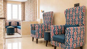 A hotel room with a seating area featuring patterned armchairs and ottomans, a sofa, and a window with curtains.
