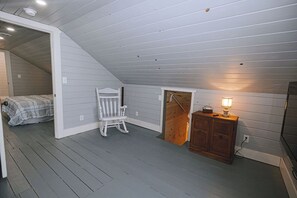 A room with a sloped ceiling, a bed with a plaid blanket, a rocking chair, a wooden cabinet with a lamp, and a door leading to another room.