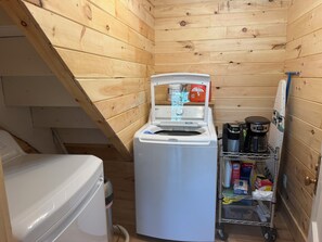 Una cabaña de madera pequeña con lavadora, cafetera y un estante con productos de limpieza.