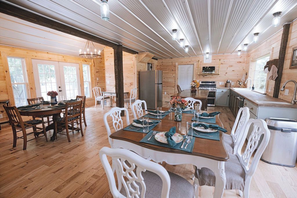 Une salle à manger spacieuse, avec une table en bois dressée pour six personnes, entourée de chaises blanches. On aperçoit, au fond, une cuisine équipée d'appareils électroménagers modernes.