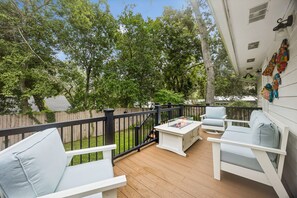 Eine Holzterrasse mit weißen Möbeln, einem schwarzen Geländer und Blick auf Bäume und einen Zaun.