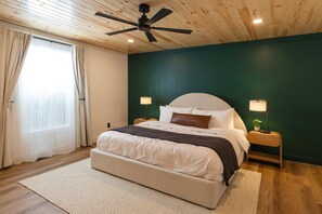 A bedroom with a bed, two bedside tables with lamps, a ceiling fan, a window with curtains, and a green accent wall. - 2 Kings | Hot Tub • Sauna • Firepit • EV • Dogs OK (Zoe)