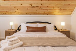 Un lit bien fait, avec des draps blancs, un oreiller marron et un plaid beige. Il y a deux tables de chevet avec des lampes, et une pile de serviettes blanches pliées.