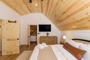 Une chambre avec un plafond en pente, des murs en bois, un lit avec une literie blanche, une commode en bois et un téléviseur à écran plat fixé au mur.