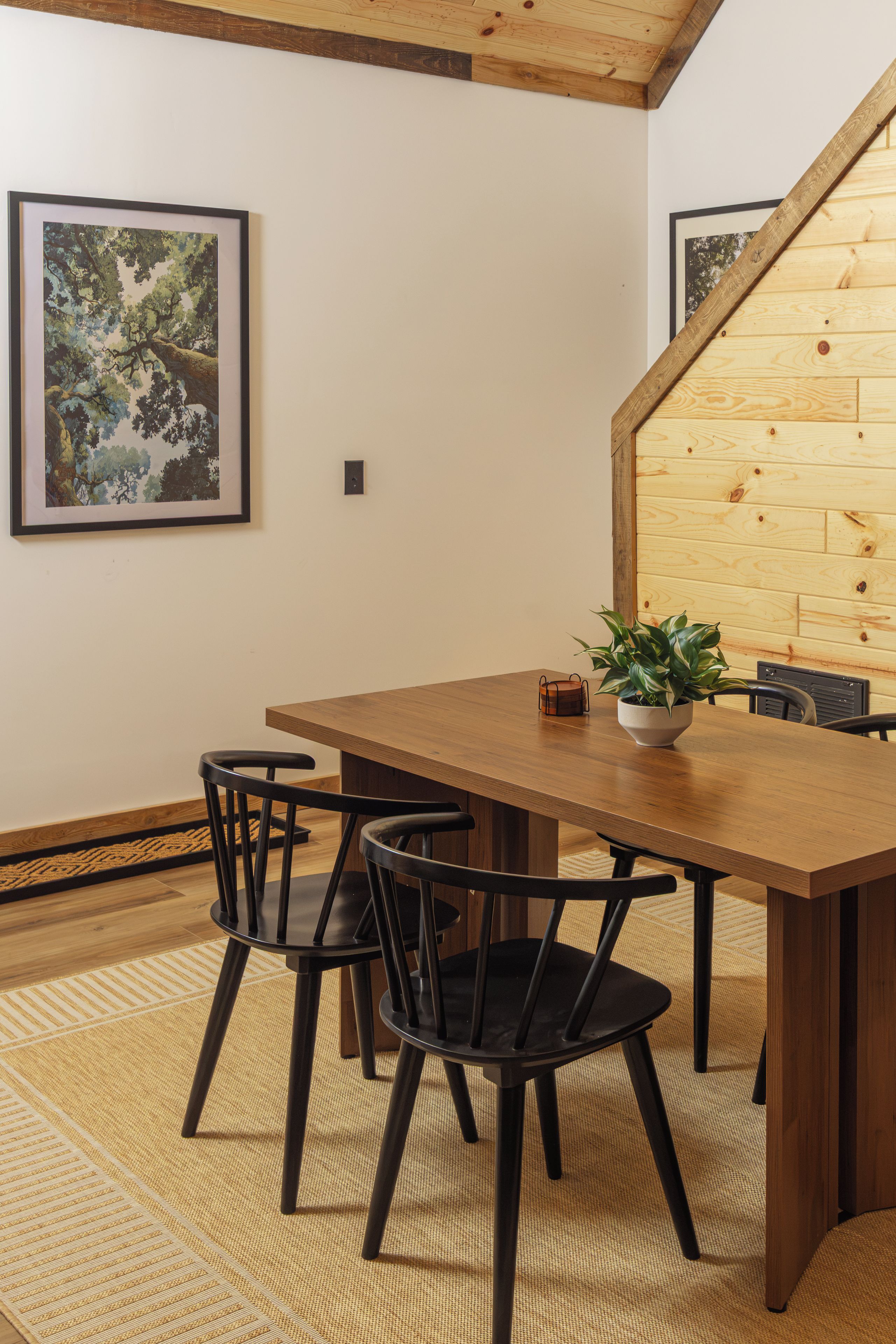 A dining area with a wooden table and chairs, a potted plant, and framed artwork on the wall.