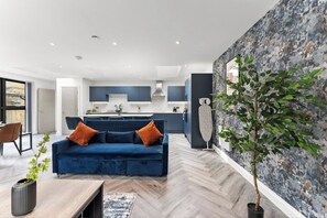 A modern living room with a blue sofa, a dining area with a table and chairs, and a kitchen in the background. - Stylish Abodes in Leeds - Sleeps 18 (Leeds)