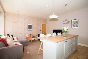 Ein modernes Wohnzimmer mit grauem Sofa, einem Holzesstisch und einer Küchenzeile mit weißen Schränken.