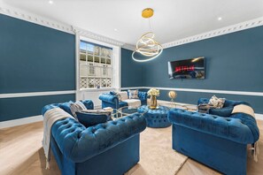 A living room with blue upholstered sofas, a coffee table, and a television mounted on the wall. - The Bath Grand | Stylish City Centre Stay for 25 (Bath)