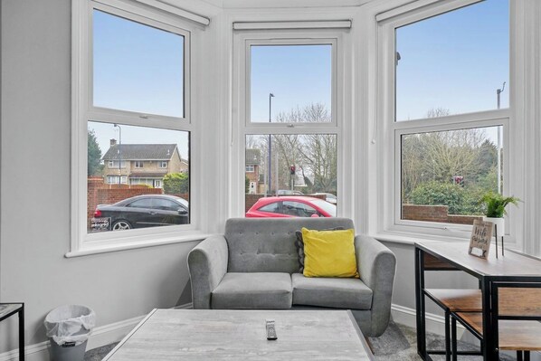 Ein Zimmer mit einem grauen Sofa, einem kleinen Tisch und einem Fenster mit Blick auf ein rotes Auto und Häuser.