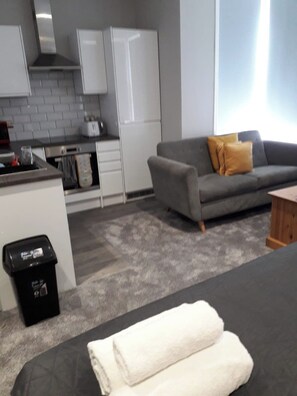 A modern living space with a kitchen area, a grey sofa with a yellow pillow, and a black trash bin. - Boutique Maidstone Gem - Sleeps 3 (Maidstone)