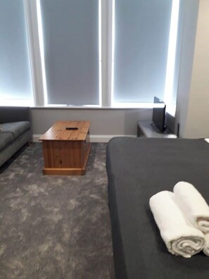 A hotel room with a bed, a wooden nightstand, a grey sofa, and a window with blinds. - Boutique Maidstone Gem - Sleeps 3 (Maidstone)