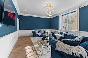 A room with a blue wall, a TV mounted on the wall, a blue sofa set, a round coffee table, and a window with a view. - Luxurious Abode in Central Bath (Bath)