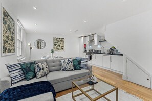 A modern living room with a grey sofa, a coffee table, and a kitchen in the background. - The Gatsby | Stylish Stay Near Roman Baths (Bath)