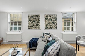 A modern living room with a grey sofa, a glass coffee table, and a vase with flowers. There are two windows with white frames and a wall-mounted artwork. - The Gatsby | Stylish Stay Near Roman Baths (Bath)