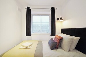 Ein kleines Zimmer mit einem Einzelbett, einem Fenster, einem Nachttisch mit einem Handtuch und einer Lampe.