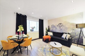 Ein modernes Wohnzimmer mit einem schwarzen Sofa, gelben Sesseln, einem Esstisch mit Blumenvase und einem großen Gemälde an der Wand.