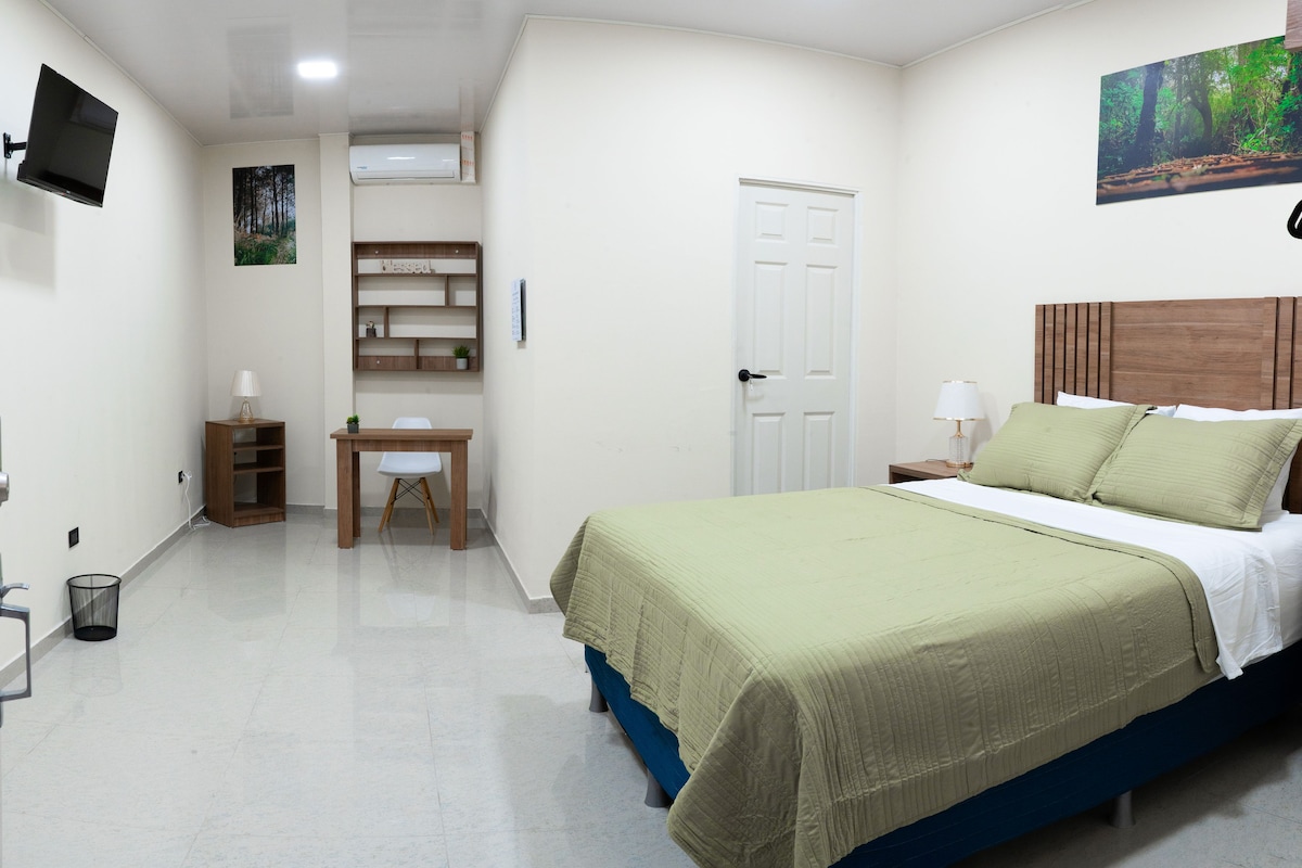 Quarto com cama, mesa de cabeceira, abajur e televisão na parede.