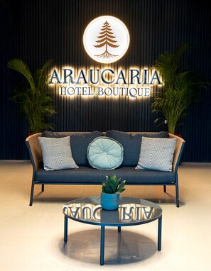 Un sofá moderno con cojines estampados, una mesa de centro de cristal con una planta, y un cartel que dice 'ARAUCARIA HOTEL BOUTIQUE' sobre un fondo oscuro.