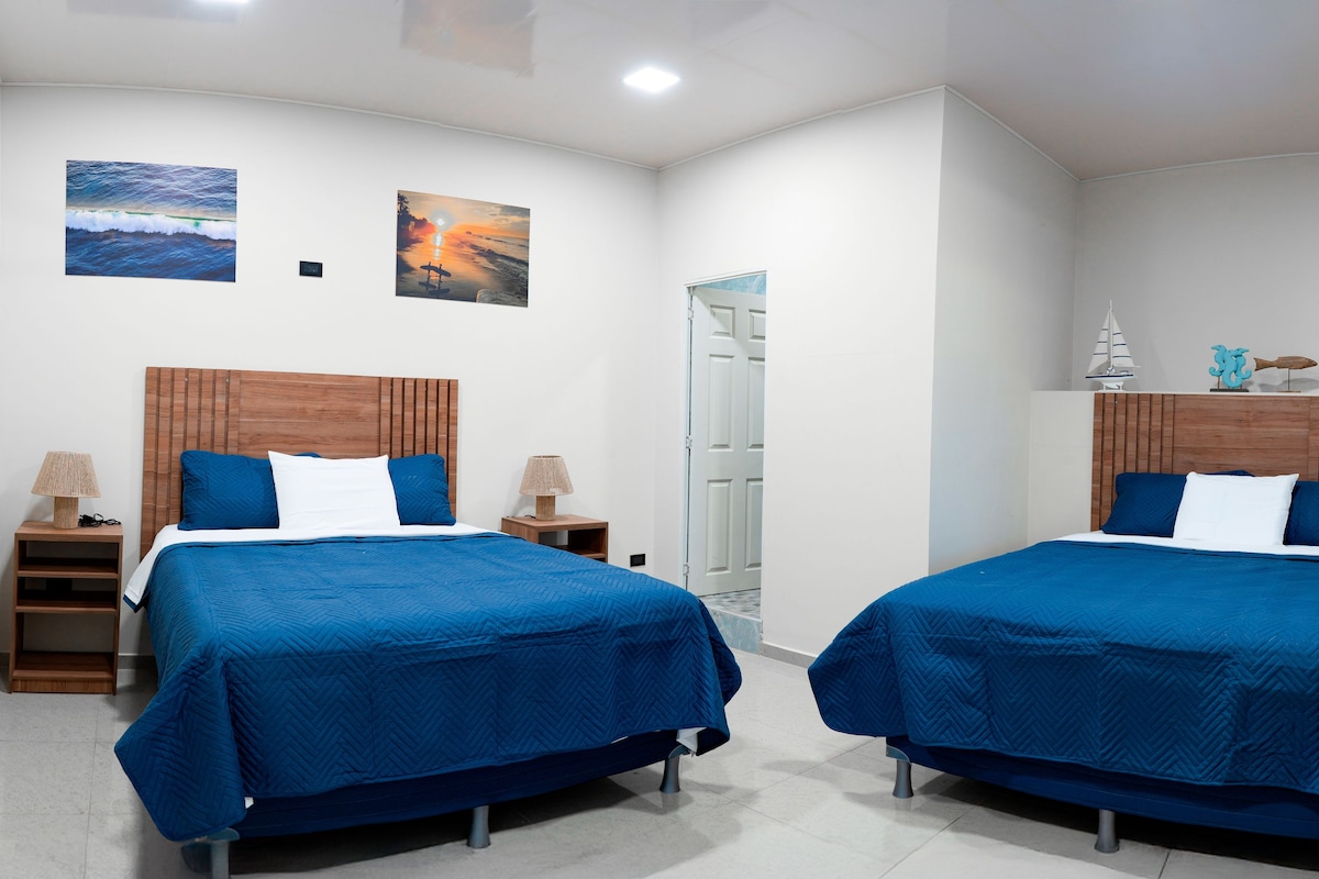 Quarto de hotel com duas camas, cabeceira de madeira, mesa de cabeceira, abajur e ar-condicionado fixado na parede.