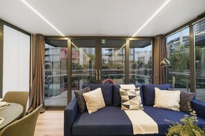 A modern hotel room with a sofa, a dining table, and a view through large glass doors.