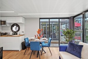 Una cocina moderna con armarios blancos, un microondas y un espejo redondo. Hay una zona de comedor con una mesa preparada para cuatro, sillas azules y vistas a un paisaje urbano a través de amplias ventanas.