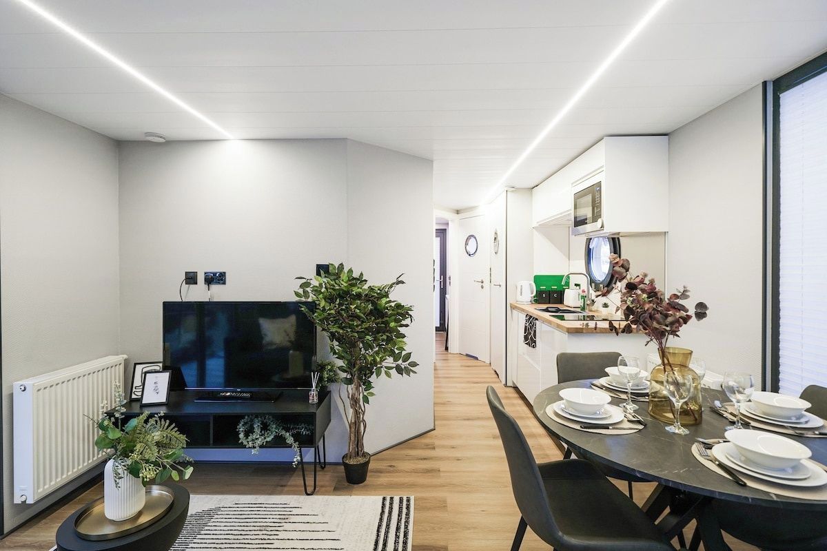 Una sala de estar moderna con un televisor de pantalla plana, una planta en maceta y una zona de comedor preparada para cuatro personas, con platos y vasos blancos.