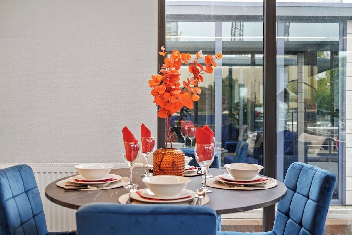 A dining table set for four with a vase of orange flowers, red napkins, and white plates.