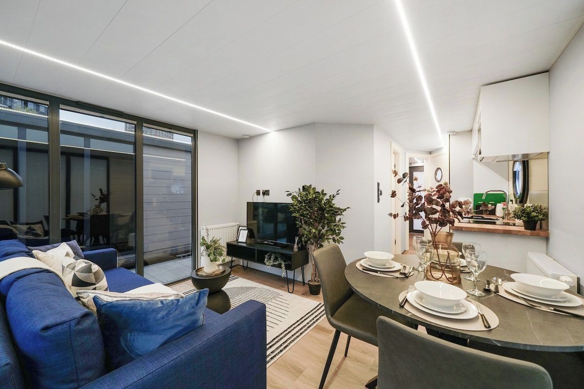 Una sala de estar moderna con un sofá, una mesa de comedor lista para cuatro personas, un televisor y un ventanal grande.