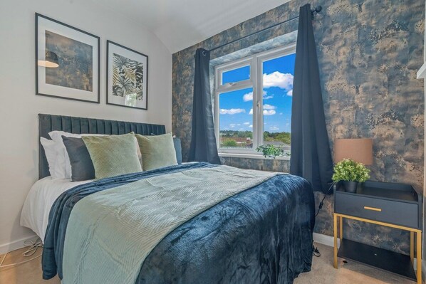 A bedroom with a bed, a window with curtains, a bedside table with a lamp, and two framed pictures on the wall. - Stylish Suburban Escape - Perivale Haven (London)