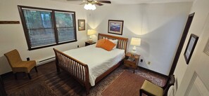 A bedroom with a wooden bed, two bedside lamps, a chair, a window with blinds, and a ceiling fan. - Secluded 2 BR Mountain Creeksong Cottage with Beautiful Fall Color Views (Burnsville)