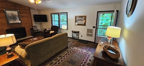 A living room with a sofa, a television, a fireplace, and a window with blinds. - Secluded 2 BR Mountain Creeksong Cottage with Beautiful Fall Color Views (Burnsville)