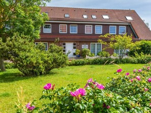 Ein zweistöckiges Haus mit rotem Ziegeldach, einer Veranda und einem gepflegten Garten mit rosa Blumen.