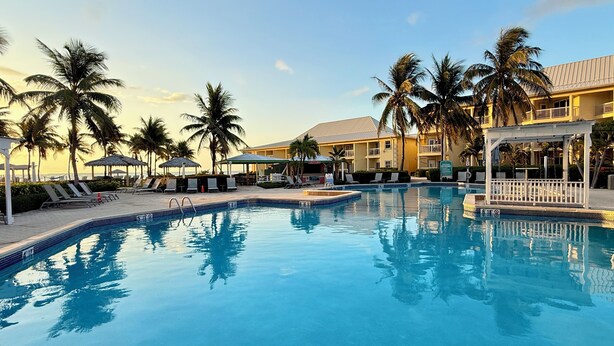 Ein Pool mit Liegestühlen und Sonnenschirmen, umgeben von Palmen und einem Gebäude mit Balkonen.