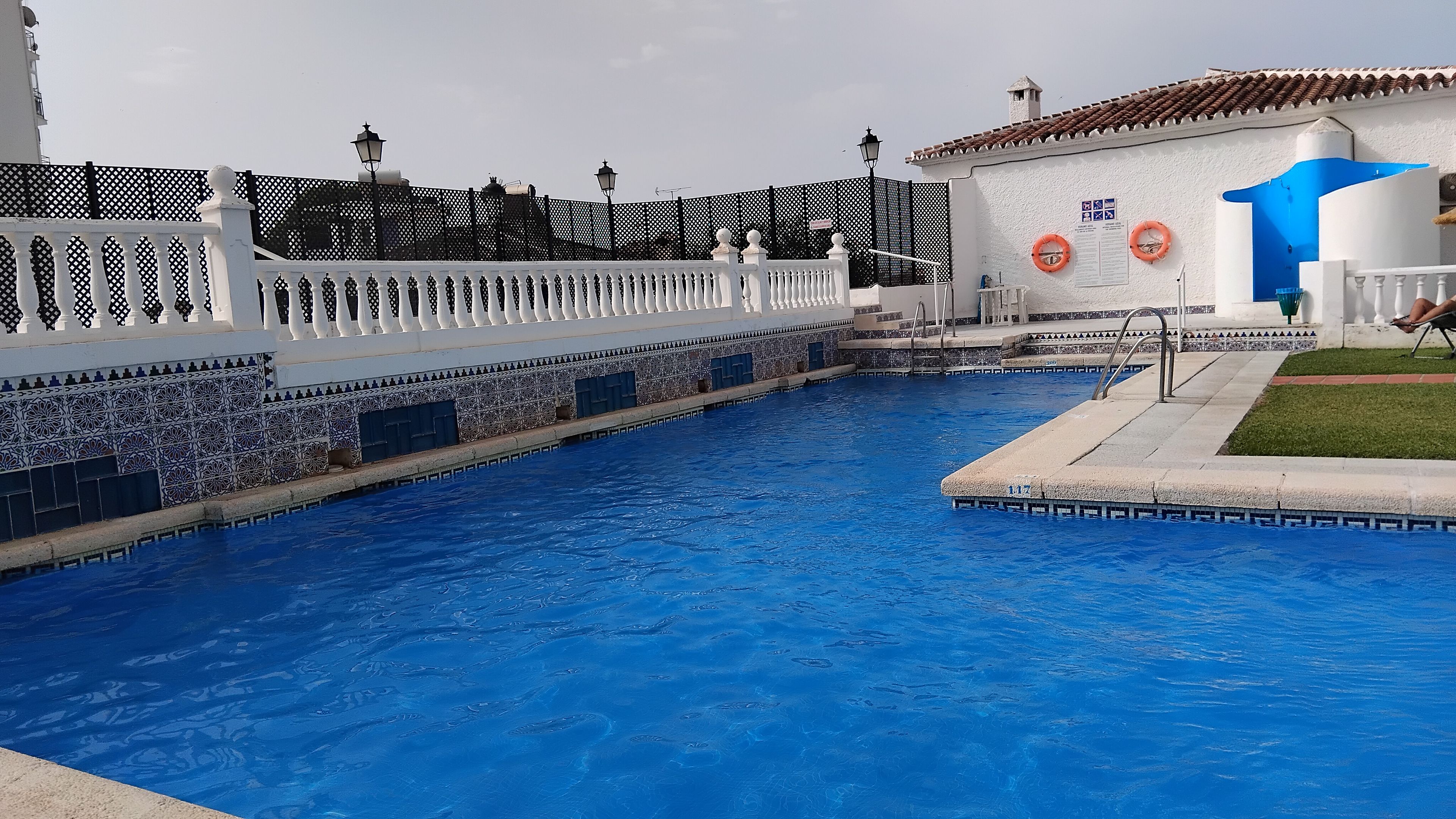 A swimming pool with a white fence, a tiled wall, and a building with a blue and white facade.