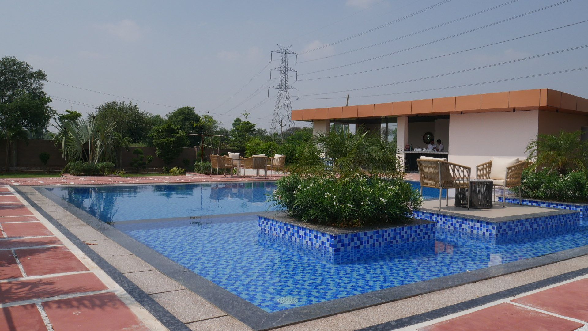 A swimming pool with blue mosaic tiles, surrounded by a brick border, and a building with a flat roof in the background.
