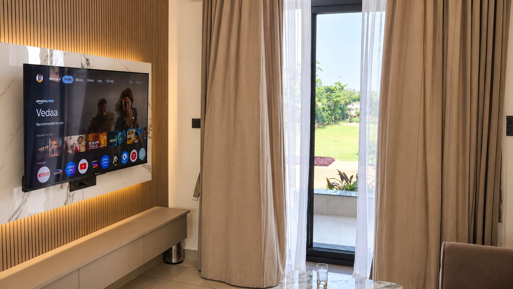 Una sala de estar moderna con un televisor de pantalla plana montado en la pared que muestra la interfaz de un servicio de streaming. Hay un banco a lo largo de la pared y vistas a un balcón a través de cortinas translúcidas.