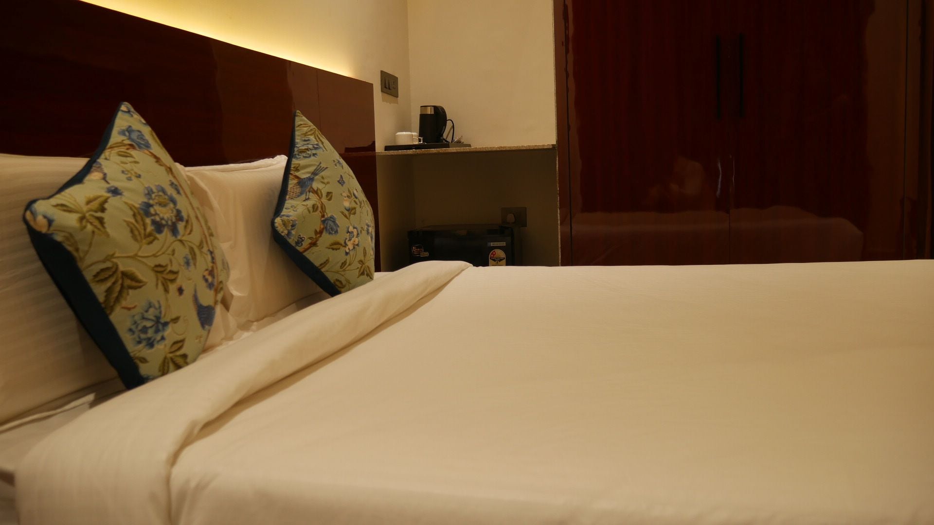 A hotel room with a bed, pillows, a coffee maker, and a shelf with a bottle.