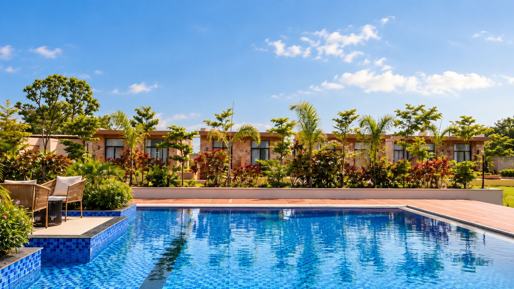 Una piscina con borde de azulejos, rodeada de exuberante vegetación y una hilera de edificios al fondo.