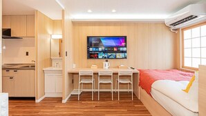 Habitación compacta con kitchenette, una televisión de pantalla plana montada en la pared, cama con manta roja y ventana con persianas.